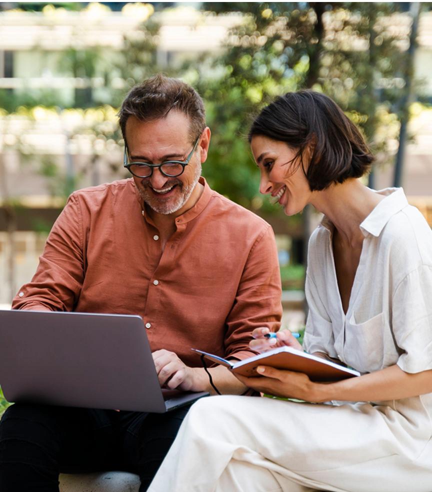 Mężczyzna i kobieta siedzą na ławce. Mężczyzna pokazuje coś na laptopie, a kobieta trzyma notatnik