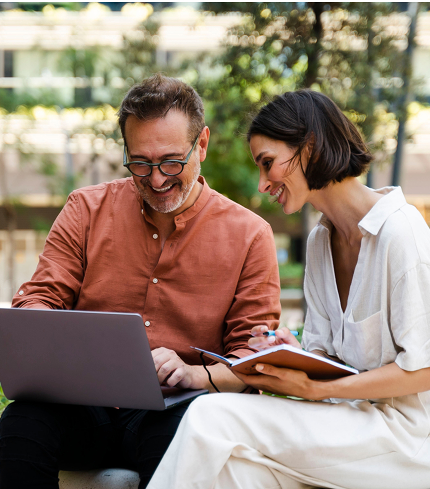 Mężczyzna i kobieta siedzą na ławce. Mężczyzna pokazuje coś na laptopie, a kobieta trzyma notatnik